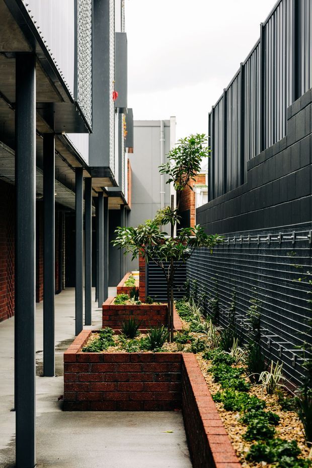 A narrow landscaped urban walkway or courtyard between contemporary buildings. The space features concrete paving, raised red-brick planter beds with small trees and drought-tolerant planting, dark metal structural posts, a black masonry boundary wall, and a tall vertically ribbed metal screen fence. The composition emphasizes strong linear perspective, layered facade materials, and compact architectural landscaping.
