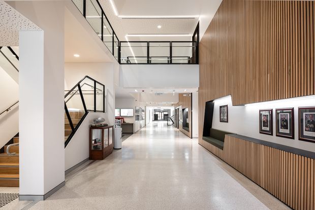 A bright, contemporary multi-level interior corridor or lobby with polished terrazzo flooring, white walls and ceilings, black-framed glass balustrades, and a stairway on the left. A tall timber batten feature wall on the right incorporates a long illuminated display niche with framed artwork and built-in dark seating. The space extends into a long open hallway beneath an upper-level void or mezzanine, with recessed and linear lighting throughout.