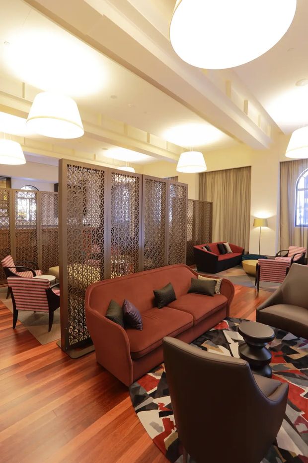 A warmly lit, richly furnished lounge or hotel-style reception area with polished timber flooring, rust-red and dark brown upholstered sofas and armchairs, a bold geometric area rug, and multiple patterned decorative screens dividing the space. Large white pendant lights hang beneath a coffered or exposed-beam ceiling, with full-height curtains and an arched window adding architectural detail. The overall atmosphere is inviting, eclectic, and refined.