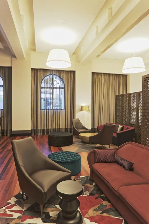 A sophisticated lounge area with tall arched windows, full-length taupe curtains, exposed cream ceiling beams, and large white pendant lights. The room is furnished with sculptural armchairs, a deep rust-red sofa, a teal tufted ottoman, round side tables, and a bold geometric patterned rug. A decorative dark metal or timber-patterned screen divides the seating area, creating a refined, warm, hotel-like atmosphere.