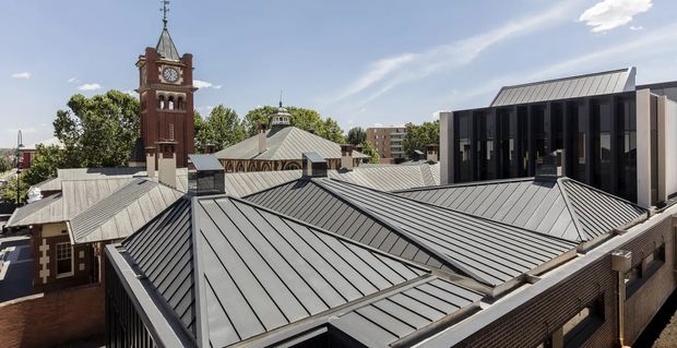 Elevated exterior view across a complex of grey standing-seam metal roofs, with a prominent historic brick clock tower and church-like roof forms integrated beside a contemporary dark-clad building. Urban surroundings and mature trees are visible in the background under a bright sky.