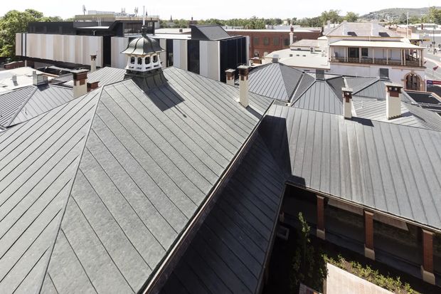 Elevated view across a dense urban rooftop landscape, dominated by grey standing-seam metal roofs with multiple pitched forms, chimneys, and a distinctive decorative cupola. Contemporary and heritage-style buildings are visible in the surrounding cityscape.