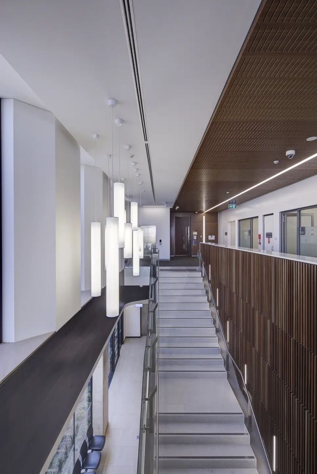A contemporary commercial interior showing a broad stairway in a bright multi-level corridor or landing. The space features a dark timber slatted ceiling and matching vertical timber balustrade, pale tiled or stone stair treads, glass and metal handrails, white walls, and a series of suspended cylindrical pendant lights. Glazed rooms line the right side, while a lower-level workspace is partially visible below.