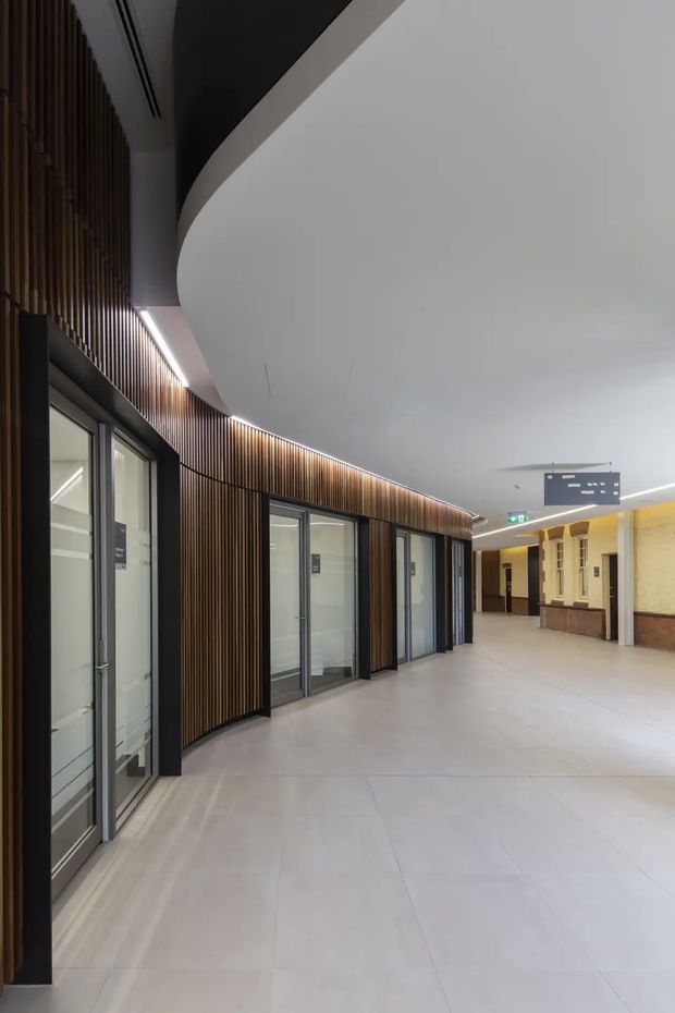 A wide modern commercial corridor with a gently curving circulation path, full-height glazed office or room doors, dark vertical timber-look slatted wall cladding, integrated linear lighting, and a smooth curved white ceiling. The corridor opens toward a brighter lobby-like area with wayfinding signage and contrasting warm-toned wall finishes.
