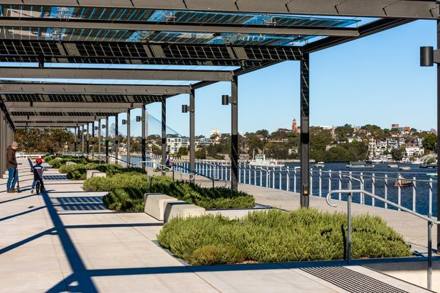 A sunny waterfront public promenade with a long black steel pergola and integrated solar-panel canopy, paved walkways, landscaped planting beds, concrete benches, lighting, and accessible railings overlooking a harbour. Urban buildings and boats are visible across the water.