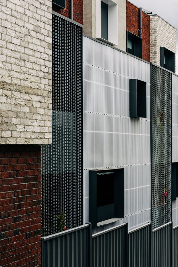 Close-up exterior view of a contemporary multi-level facade composed of pale stone masonry, red brickwork, translucent ribbed panel cladding, dark metal window surrounds, and vertical chain-screen elements. Dark corrugated metal fencing and recessed openings reinforce the industrial-modern character. No interior room is visible.
