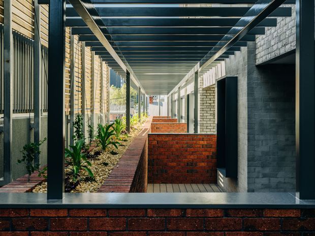 A long, covered architectural passageway with strong linear perspective. Dark steel posts and overhead beams frame a narrow landscaped strip with low planting and brick-edged paths. Red brick privacy walls and planters contrast with dark painted masonry walls, vertical timber screening, and pale brickwork. The space appears to connect multiple building entries or wings and is photographed in bright daylight.