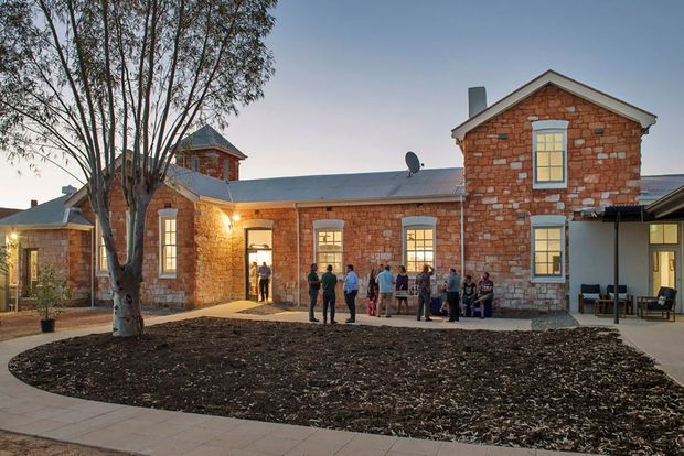 Exterior view of a restored red-brick heritage building arranged around a landscaped courtyard. The single-storey wing has a low metal roof, while the taller gabled section features stone detailing, tall timber-framed windows, and a chimney. People gather near the illuminated entrance at dusk, with paved paths and a central garden bed in the foreground.