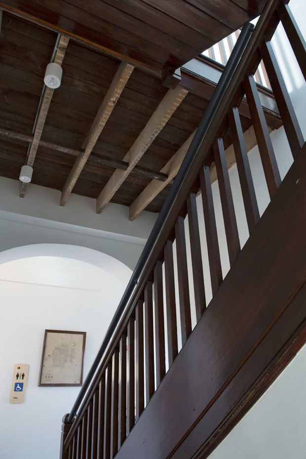 Interior view looking upward along a dark timber staircase with vertical balustrades and a dark handrail. The stair rises beneath an exposed timber ceiling with visible beams and rafters, while white arched walls and clerestory-style glazing bring light into the circulation space. A framed artwork and accessibility signage are visible on the wall.