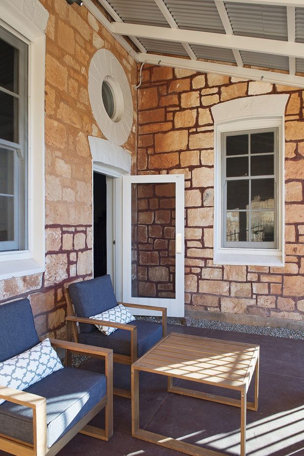 A covered outdoor veranda or porch with distinctive exposed sandstone masonry walls, white-painted timber-framed windows and door, and a corrugated metal roof supported by white beams. Two blue upholstered timber armchairs with patterned cushions face a slatted timber coffee table on a dark paved floor. Sunlight casts strong linear shadows across the seating area, creating a relaxed heritage character.