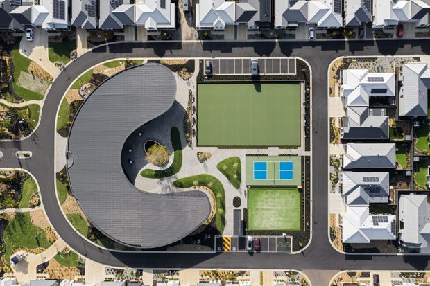 Top-down aerial view of a planned residential development centred around a large organic, curved-roof community or recreation building. The site includes a full-size rectangular sports court or field, two smaller blue courts, landscaped pedestrian paths, parking areas, internal roads, and tightly arranged detached homes with light-coloured pitched roofs.