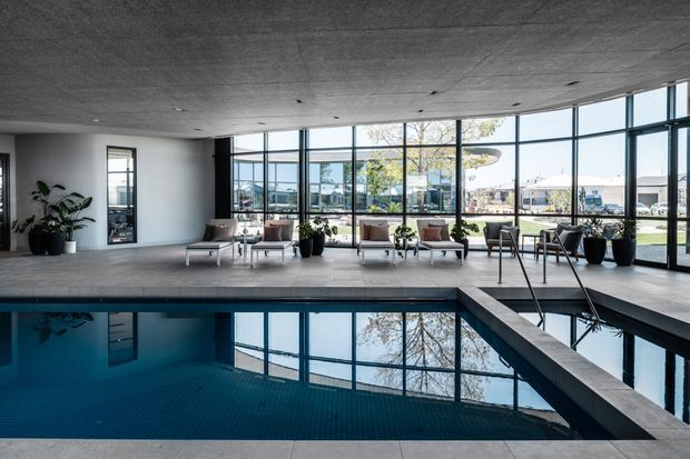 A contemporary indoor pool lounge with a dark tiled swimming pool in the foreground, surrounded by pale stone or concrete paving. Floor-to-ceiling black-framed glazing overlooks an outdoor setting, while a relaxed seating area with armchairs, lounge chairs, coffee tables, and potted plants lines the perimeter. The space has a low exposed concrete ceiling with recessed downlights and a calm, refined atmosphere.