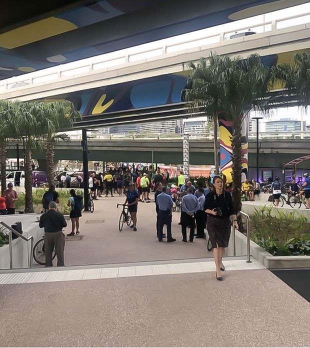 A busy open-air commercial transit or civic plaza beneath elevated pedestrian bridges and covered structures. The space features broad paved walkways, stairs with metal handrails, palm trees in raised planters, cyclists, pedestrians, and colorful graphic panels integrated into the overhead architecture.