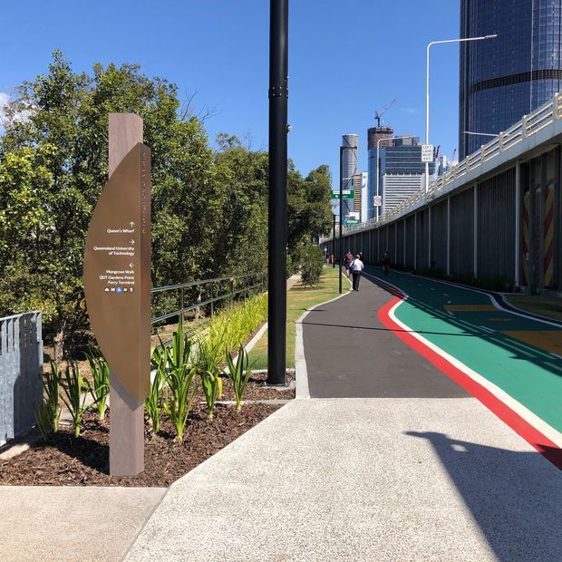 A sunny urban pedestrian walkway beside a large elevated roadway or bridge, with colourful painted path markings, landscaped planting, railing, and a tall directional wayfinding sign. Modern city high-rise buildings are visible in the background.