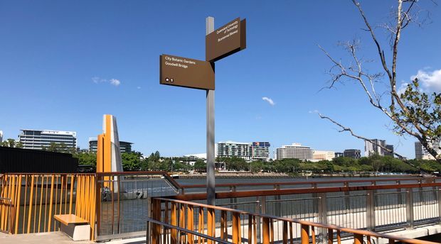 Outdoor pedestrian waterfront or urban park walkway with orange-painted railings, timber handrails, metal balustrades, and directional signage. Modern mid-rise buildings and landscaped public space are visible beyond the railing under a clear blue sky.