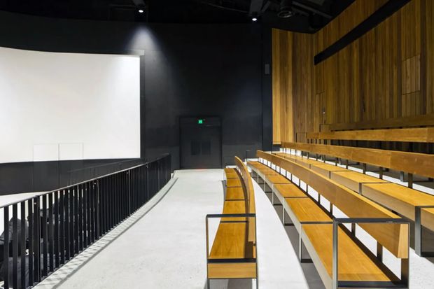 A modern tiered auditorium or lecture theatre viewed from the side aisle, with long rows of fixed timber bench seating, slim black metal supports and railings, a large projection screen, dark acoustic walls and ceiling, and a timber-paneled feature wall. The space is clean, minimal, and suited to presentations or performances.