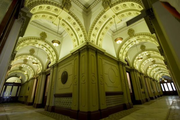 Interior view of an ornate historic corridor or public hall with repeated barrel-vaulted arches, elaborate decorative plasterwork, classical motifs, pendant chandeliers, paneled pilasters, and polished stone flooring. The symmetrical perspective and richly embellished ceiling create a grand, heritage character.