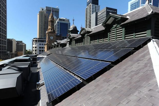 Exterior rooftop view of a heritage-style building in a dense city centre, dominated by a broad sloping tiled roof fitted with rows of photovoltaic solar panels. The roof includes dark timber louvred or weatherboarded elements, ornate gables and a small turret-like feature, with modern high-rise office buildings visible in the background under a clear blue sky.