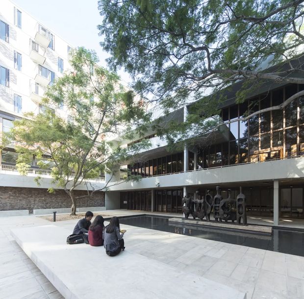A bright modern institutional courtyard framed by a multi-storey white apartment or academic building on the left and a low, dark-glazed concrete building on the right. The open plaza has pale stone paving, a long shallow reflecting pool, mature trees, and a dark sculptural installation beneath the glazed colonnade. Several people sit on a raised platform in the foreground.
