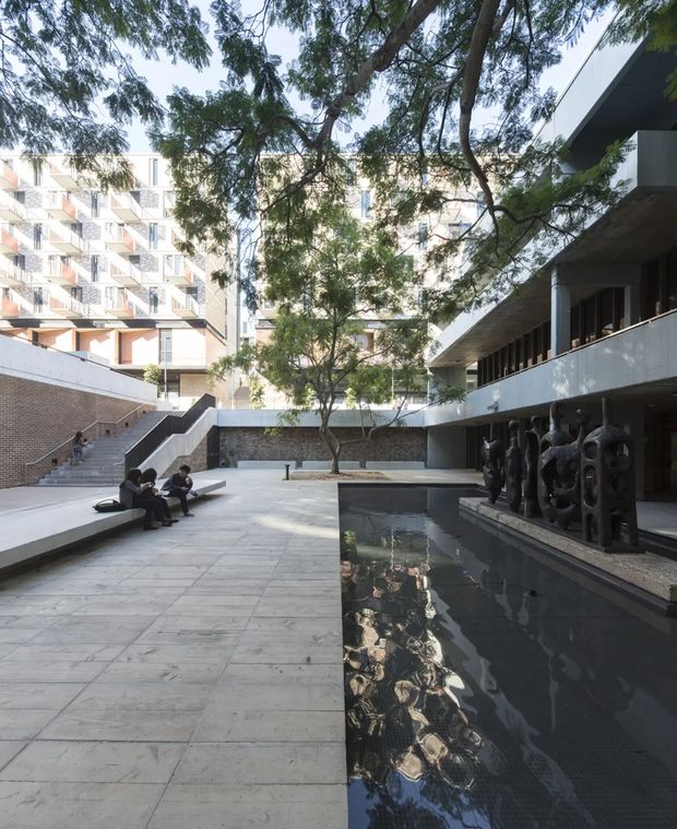 A spacious modern institutional courtyard framed by multi-storey buildings, mature trees, broad concrete paving, stepped seating, and a long shallow reflecting pool. A covered walkway with dark sculptural figures lines the right side, while apartment-like facades with repetitive balconies rise in the background. The scene has a calm, urban architectural character with strong geometric forms and filtered natural light.
