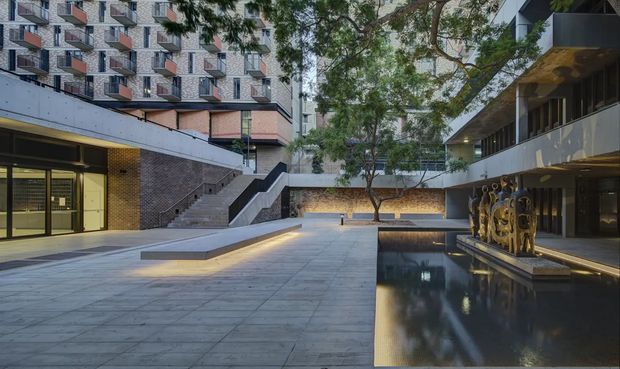 A landscaped urban courtyard or forecourt within a multi-storey contemporary complex. Broad pale paved surfaces lead to stepped terraces and lower-level entries, with dark brick and concrete walls, recessed linear lighting, mature trees, and a long reflective water feature beside a covered walkway. Apartment or institutional-style buildings surround the open space, with repetitive balconies and sculptural artwork along the pool edge.