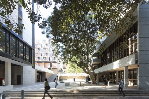 A tree-lined urban pedestrian courtyard framed by contemporary multi-storey buildings. Broad concrete steps and paved open space connect the buildings, with extensive ground-floor glazing, dark window frames, pale concrete or rendered facades, and people moving through the plaza. The scene has a bright, spacious institutional or commercial character.