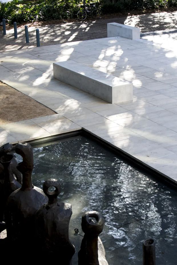 A landscaped outdoor courtyard or plaza with a shallow rectangular water feature in the foreground, including dark sculptural fountain elements. The surrounding area is paved in large pale square tiles and incorporates simple integrated concrete bench walls. Dense planting, bicycle racks, and dappled tree shade appear in the background. The scene has a restrained contemporary civic or commercial landscape design.