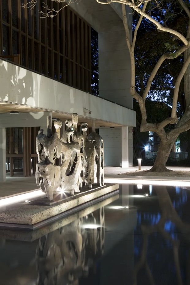 Night-time exterior view of a modern building courtyard with deep overhanging concrete forms, dark vertical window screening, and a shallow reflecting pool. A sculptural stone or concrete water feature sits on a raised plinth in the foreground, with integrated linear lighting and mature trees surrounding the space.
