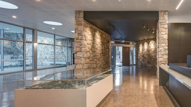 A spacious contemporary interior lobby or entry area featuring exposed rustic stone walls and columns, polished stone or terrazzo flooring, extensive floor-to-ceiling glazing, recessed and track lighting, and a large stone-topped display or reception island. Dark ceiling panels contrast with the warm stonework and pale joinery.