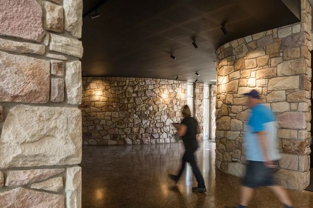 A dimly lit architectural interior with substantial rough-faced stone walls and rounded stone-clad columns. The polished dark floor reflects warm spot lighting, while a dark ceiling with track lights spans the open circulation space. Blurred people are walking through the space, suggesting a public or commercial interior such as a gallery, visitor centre, or lobby.