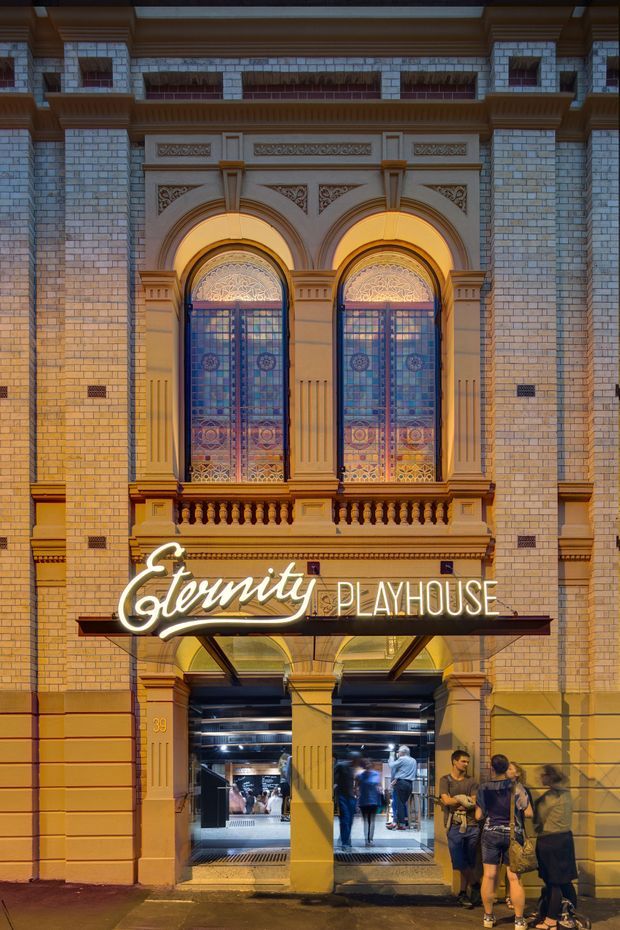 Street-level night view of the ornate Eternity Playhouse theatre facade. The historic masonry frontage features pale brick walls, elaborate classical decorative mouldings, arched stained-glass windows, a small balustrade, illuminated marquee signage, and a glazed entrance with people visible inside.