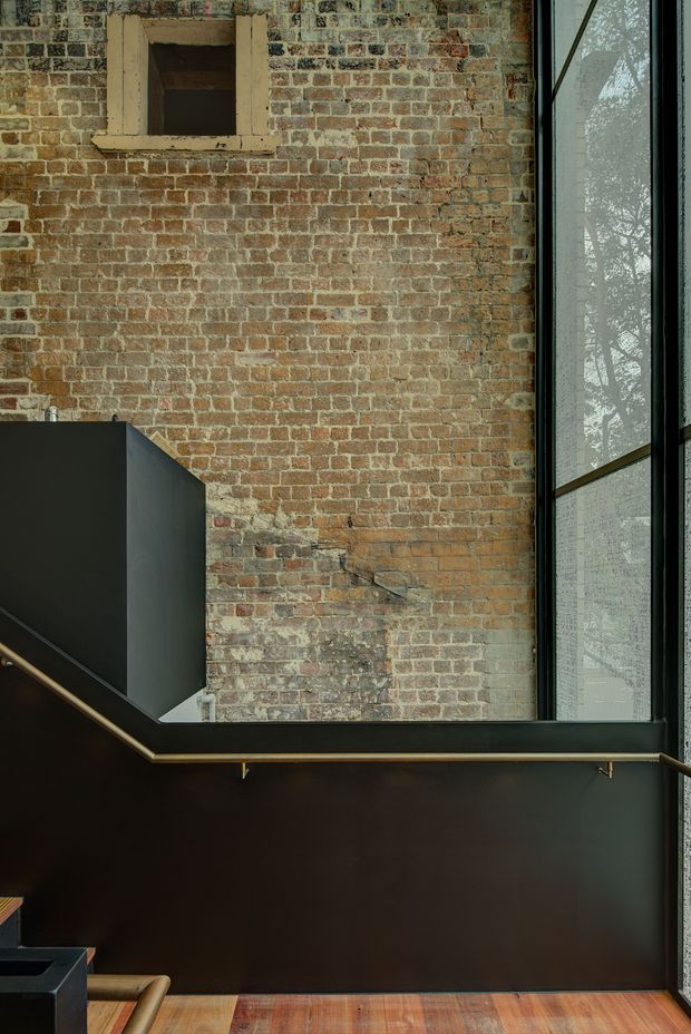Interior architectural detail featuring an exposed aged brick wall, a tall black-framed glazed opening, and a dark angular stair or balustrade structure with a slender brass-toned handrail. Warm timber flooring and dramatic vertical glazing create an industrial, adaptive-reuse atmosphere.