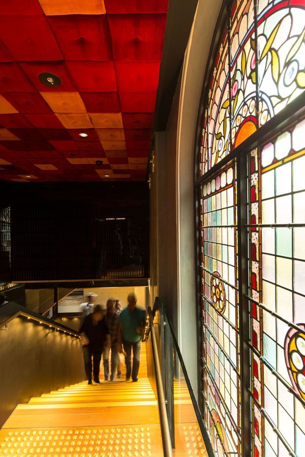 Interior architectural photograph of a dramatic stairway in a dark, multi-level public or commercial space. Warm timber stair treads and handrails are illuminated with concealed lighting, while a large ornate stained-glass window dominates the right side. The ceiling features a grid of richly coloured red, orange, and wood-toned panels with recessed spotlights. Several people are walking on the stairs, providing scale and movement.