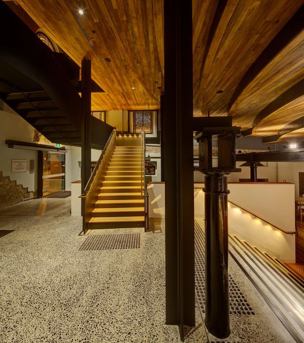 Interior view of a dramatic multi-level stairway and landing in a converted industrial or heritage building. The space features exposed dark steel structural columns and pipes, a warm timber-lined ceiling with recessed lighting, illuminated stair treads, terrazzo flooring, and a second descending stair visible to the right. Original masonry and industrial details remain visible around the circulation space.