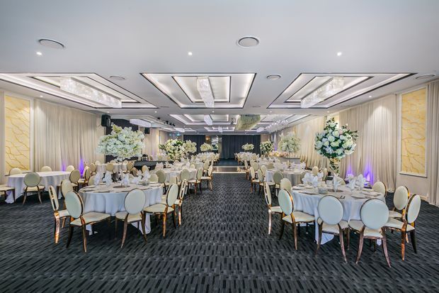 A large, symmetrical banquet or event hall arranged for a formal reception, with numerous round tables dressed in white linens, upholstered dining chairs, floral centrepieces, and a stage or focal area at the far end. The room features patterned dark carpet, cream drapery and wall panels, elaborate recessed ceiling coffers with integrated lighting, chandeliers, and decorative floral displays.