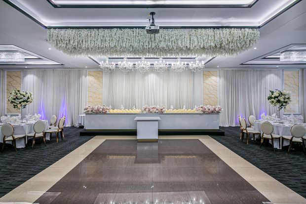 Formal, symmetrical ballroom or event hall with a central raised stage or head table, white draped walls, floral arrangements, crystal chandeliers, dramatic cascading ceiling installation, and a polished geometric stone floor. The space has an upscale wedding or banquet setting with cool white and soft purple accent lighting.