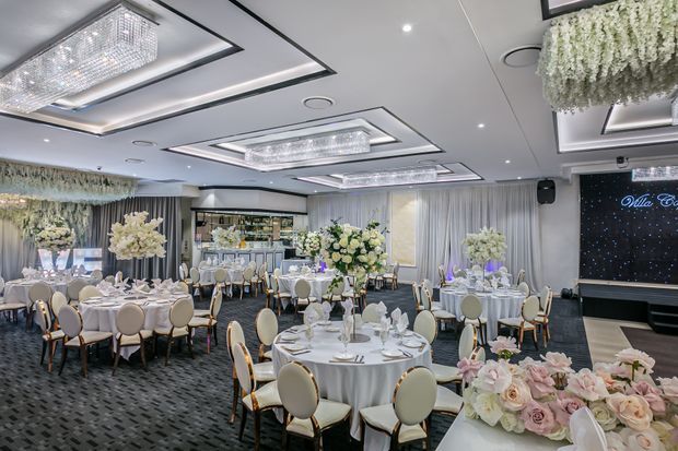 A luxurious formal event or wedding reception hall with numerous round tables dressed in white linens, elegant upholstered chairs, floral centrepieces, crystal chandeliers, layered recessed ceiling details, draped walls, and a dark feature stage or screen at the rear. The room has a polished, ornate contemporary banquet-hall aesthetic.