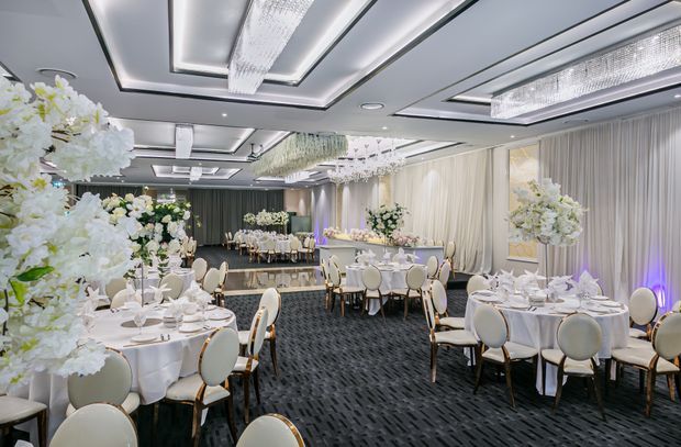 A large, elegant banquet or event hall arranged with multiple round dining tables, white tablecloths, place settings, upholstered chairs, and abundant white floral arrangements. The room features dark patterned carpet, full-height drapery, mirrored or reflective wall panels, and layered recessed ceiling coffers with long crystal-style pendant fixtures. Cool white lighting with subtle purple accent illumination creates a refined formal atmosphere.