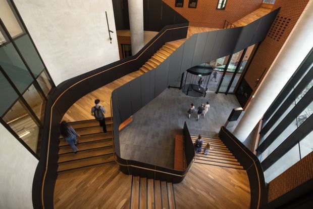 A dramatic multi-level interior atrium with sweeping sculptural timber staircases and dark curved balustrades wrapping around a central open landing. The space features warm timber stair treads and flooring, exposed red brick walls, pale plaster surfaces, extensive black-framed glazing, and a tall cylindrical structural column. Several people provide scale, while a small round table and chairs occupy the central landing.
