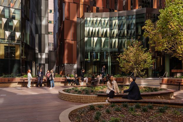 A landscaped urban courtyard or public plaza framed by tall contemporary commercial buildings. The space features curved brick-edged raised garden beds with integrated timber bench seating, mature trees, paved walkways, and groups of people using the outdoor seating. The surrounding facades combine dark metal or glass surfaces, warm reddish cladding, and striking patterned glazing.