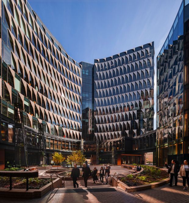 A contemporary urban commercial courtyard enclosed by several mid- to high-rise office buildings. The facades feature extensive dark glazing and repetitive projecting triangular sun-shading fins, creating a strong geometric pattern. Landscaped raised garden beds, brick-paved walkways, seating, stairs, and pedestrians occupy the central public plaza. The image is photographed outdoors in clear daylight and emphasizes the architecture and shared urban space.
