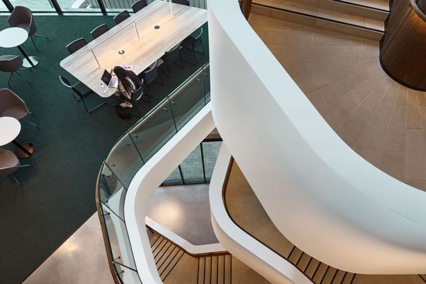An elevated interior view of a contemporary multi-level commercial or institutional space, centered on a sweeping white curved staircase and balustrade. The stair connects several levels around an open void, with timber stair treads, glass balustrades, pale tiled flooring, and a visible meeting or dining area with a long table and chairs below. Large glazed windows bring in daylight, creating a refined, airy architectural composition.