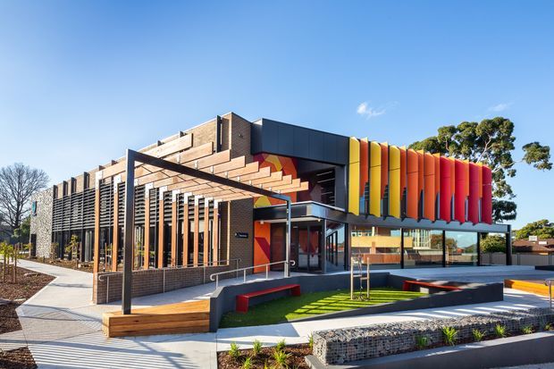 Exterior photograph of a contemporary multi-storey public or commercial building with a strongly articulated facade. Dark brick walls, projecting horizontal brick bands, timber-toned vertical screens, black metal framing, extensive glazing, and a vivid row of yellow, orange, and red vertical facade fins define the design. The landscaped forecourt includes paved paths, raised seating or planter edges, and young trees under a clear sky.