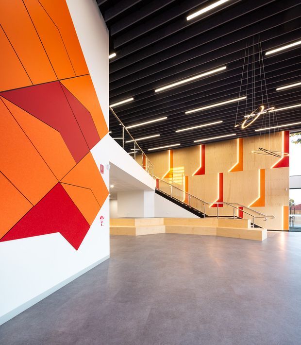 A spacious contemporary institutional or commercial interior lobby with a dramatic orange and red geometric wall mural, exposed dark ceiling structure, linear suspended lighting, warm timber wall panels with integrated angular orange accents, and a broad polished grey floor. A stair and mezzanine-style circulation route occupy the rear, with open glazing at the right.