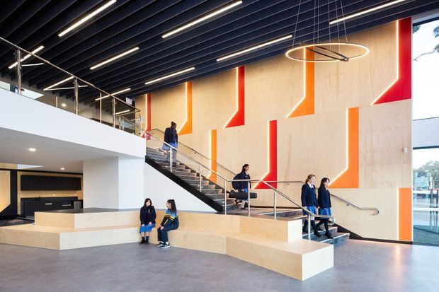 A spacious contemporary multi-level interior, likely an institutional or commercial atrium, with a broad stairway and integrated pale timber bleacher-style seating. The dramatic wall behind the stairs features geometric orange and red painted panels over light timber, while a dark linear ceiling contains long recessed strip lights and a circular pendant fixture. Large glazing on the right brings in daylight, and several people provide scale.