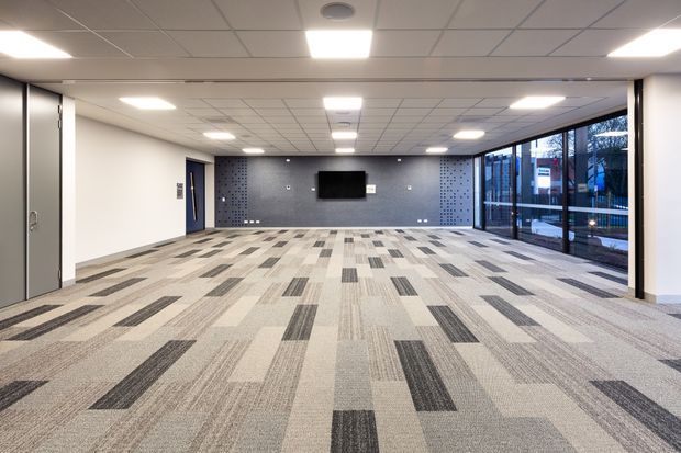 A large, open-plan commercial interior with a broad carpeted floor, white walls, a suspended acoustic tile ceiling with recessed and panel lighting, and a dark patterned feature wall at the far end with a wall-mounted screen. Full-height glazing along the right side provides views outside. The space appears unfinished or minimally furnished, suitable for a meeting, event, office, or multipurpose area.