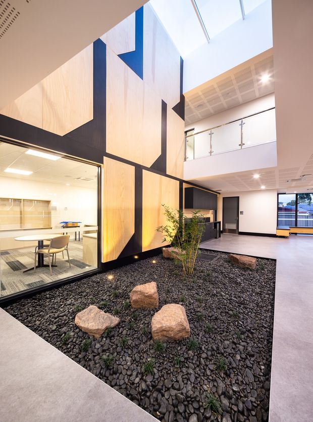 A bright contemporary commercial interior centred on a landscaped indoor courtyard filled with dark rounded stones, scattered natural boulders and a small planted tree. The surrounding space features dramatic geometric light timber and dark wall panels, extensive glazing into a meeting or dining area, an upper-level balcony with glass balustrade, skylights, and exposed services. Neutral polished flooring and white ceilings create an airy, modern atrium-like environment.