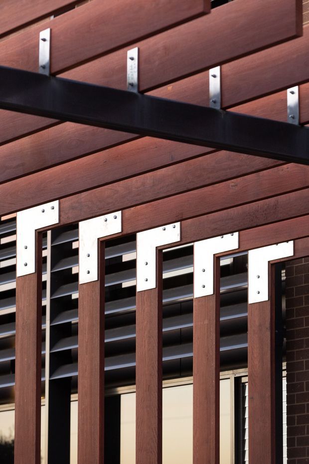 Close-up architectural detail of a contemporary exterior facade featuring dark reddish-brown timber slats and vertical timber framing, connected with exposed silver metal brackets and fasteners. Black steel beams and horizontal louvres create a strong geometric composition, with a dark brick wall visible at the side.