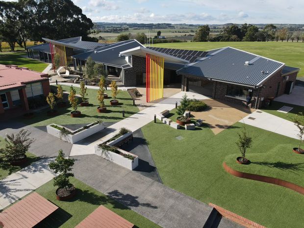 Aerial view of a low-rise contemporary institutional or commercial building arranged around landscaped courtyards and paved walkways. The building has dark grey metal pitched roofs, dark brick walls, extensive glazing, and a prominent vertical multicoloured slatted feature in yellow, orange, and red. Surrounding lawns, geometric garden beds, seating areas, and mature trees are visible, with open rural farmland beyond.
