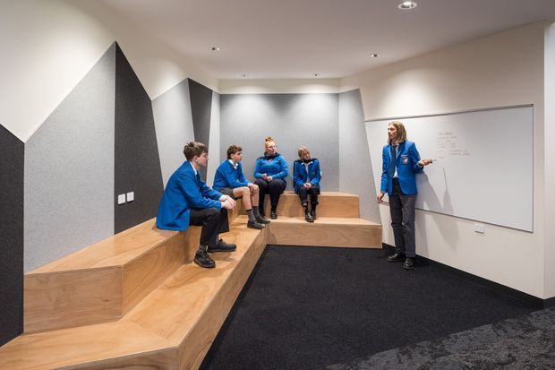 A contemporary collaborative meeting and presentation space with tiered built-in timber seating, dark carpet, acoustic wall panels, recessed ceiling lights, and a large whiteboard. Several people in blue uniforms are seated on the stepped platform while another presents beside the whiteboard.